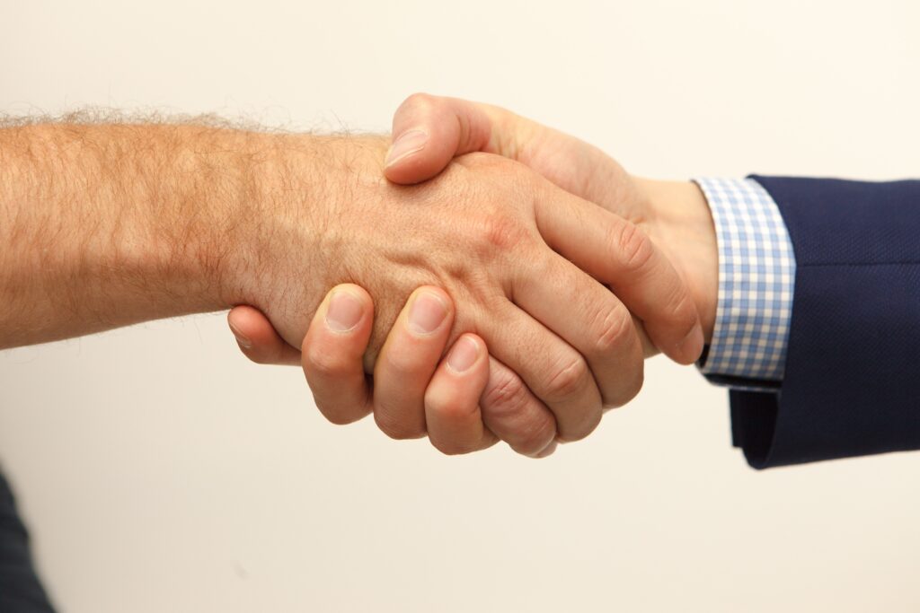 An insurance agent engaging in a firm handshake with a client, one with a bare arm and the other wearing a blue suit sleeve.